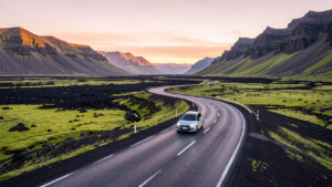 Voiture de location roulant au lever du jour sur une route islandaise entre des montagnes volcaniques et une végétation de mousse verte.
