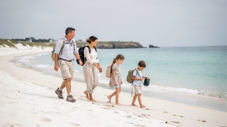 Un homme, une femme, une jeune fille et un jeune garçon marchent ensemble sur une plage de sable blanc à Houat, avec de légers sacs et la mer turquoise sous une douce lumière bretonne.