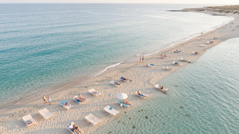 Vue aérienne réaliste d’une plage méditerranéenne en septembre, avec une eau turquoise, des transats espacés et quelques voyageurs discrets dans la lumière douce de fin d’été.